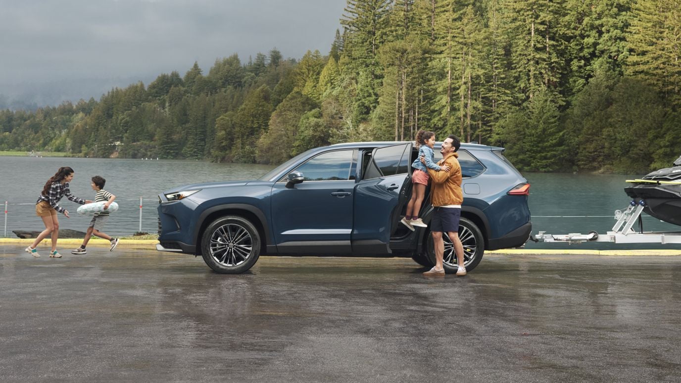 A blue Toyota Grand Highlander in profile, parked by a lake, with a family playing around it
