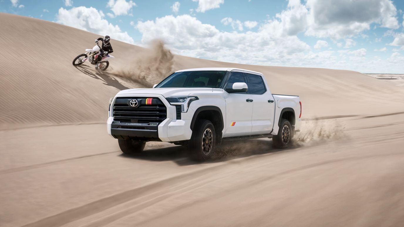 A white 2026 Toyota Tundra driving over sand with a dirt bike driving on a dune beside it