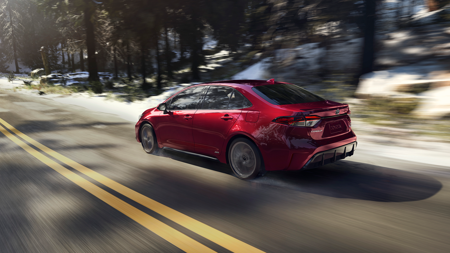A red Toyota Corolla driving down the road by a slightly snowy forest