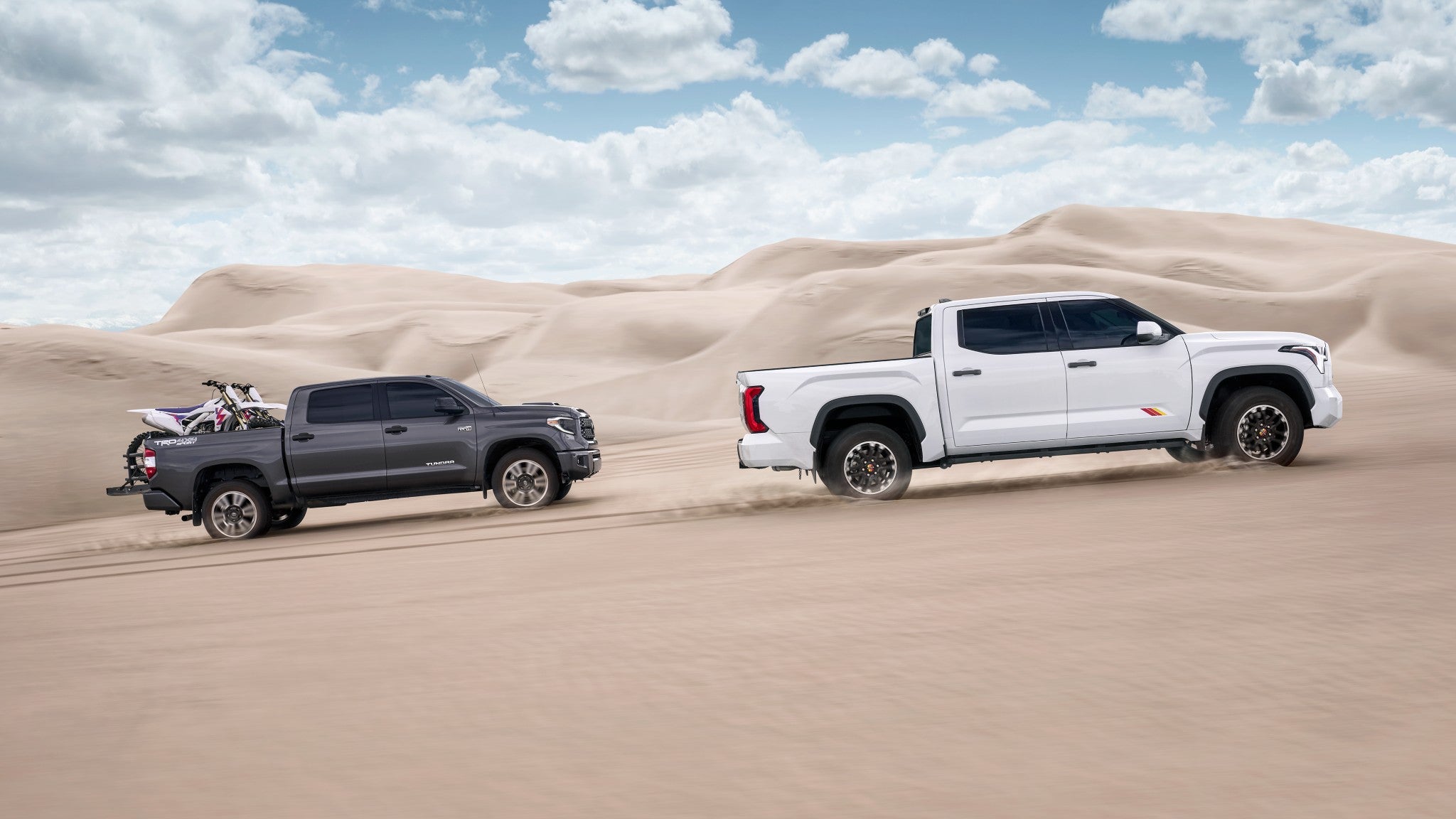 A white and grey Toyota Tundra driving over sand dunes, with the grey Tundra hauling dirt bikes