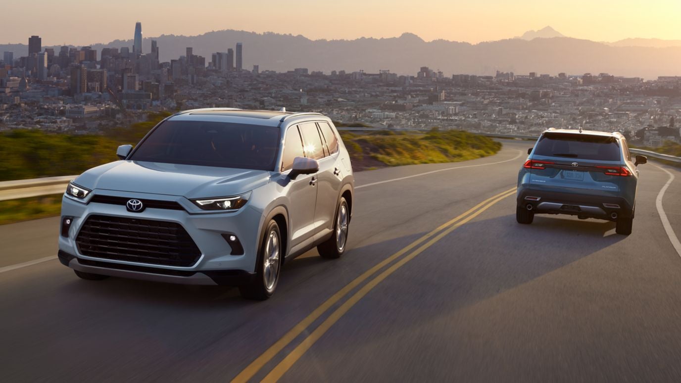 A silver and blue Toyota Grand Highlander passing each other on the road with a city in the background