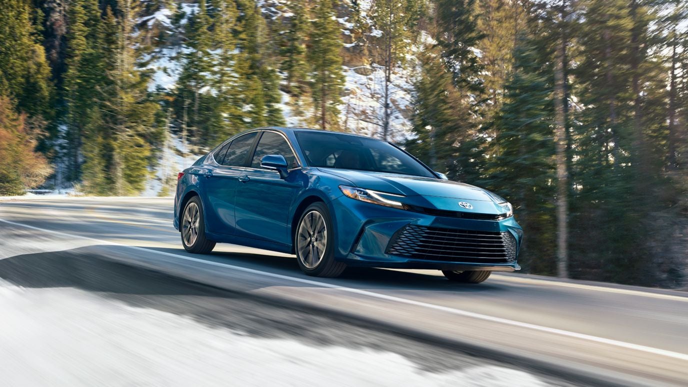 A blue Toyota Camry driving down the road by a snowy hillside and forest